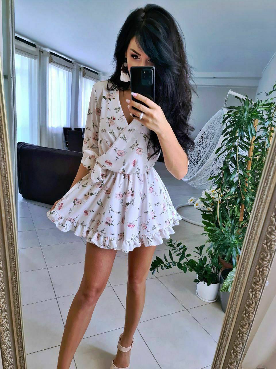 Woman taking a mirror selfie wearing a floral dress in a room with plants.