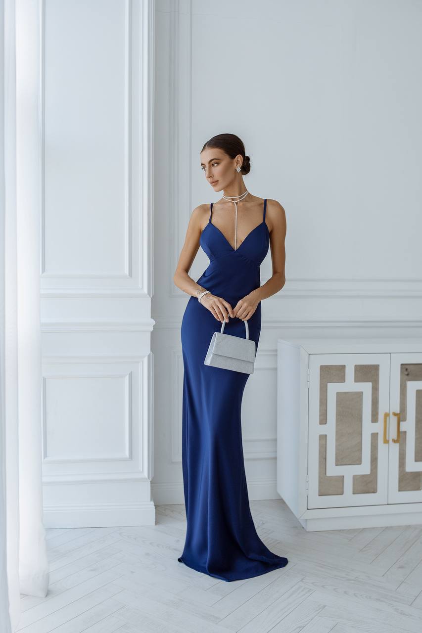 Woman in a blue evening gown holding a white handbag in an elegant room.
