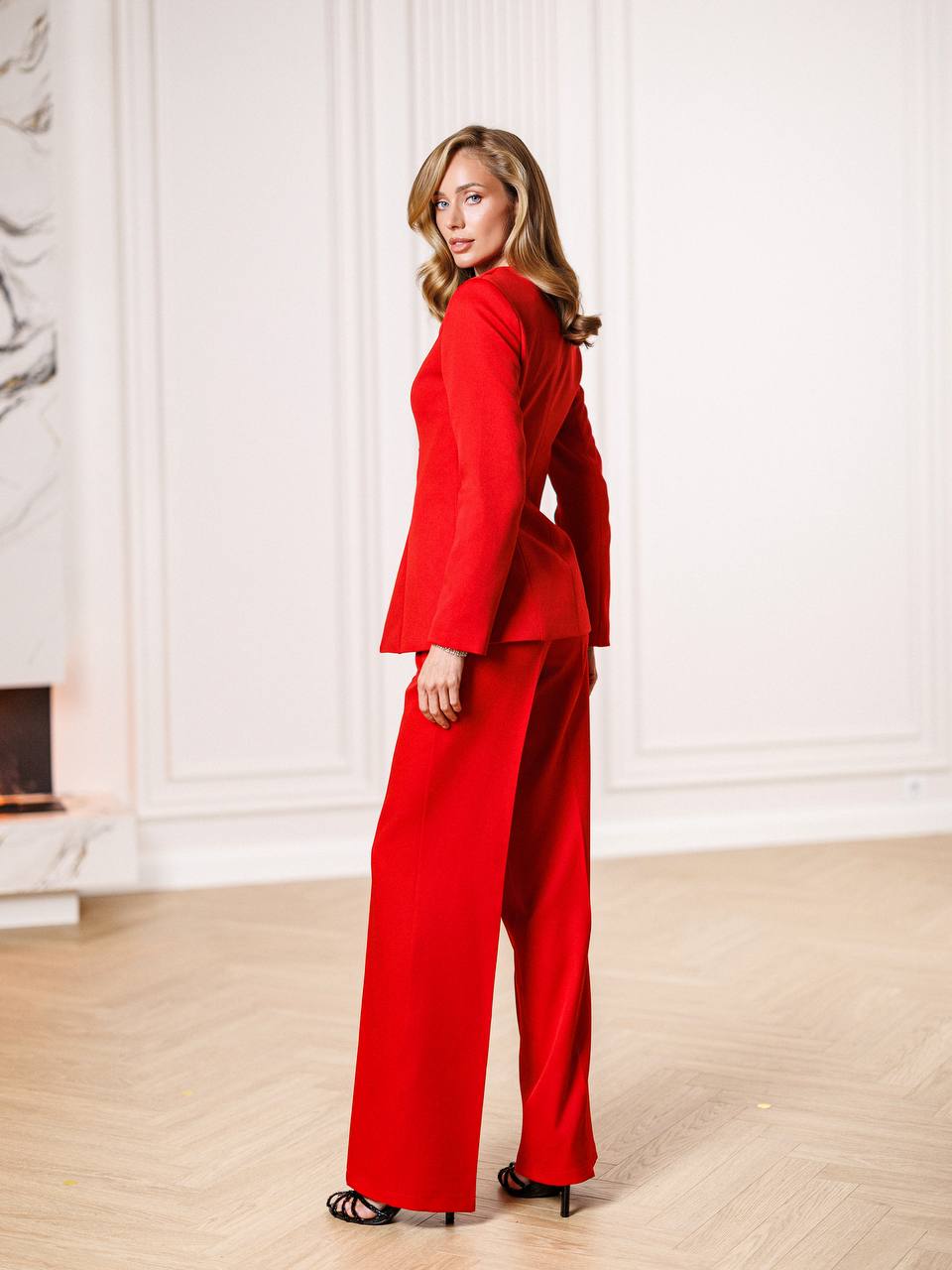 Woman wearing a red suit standing in a room with white walls and wooden floor.