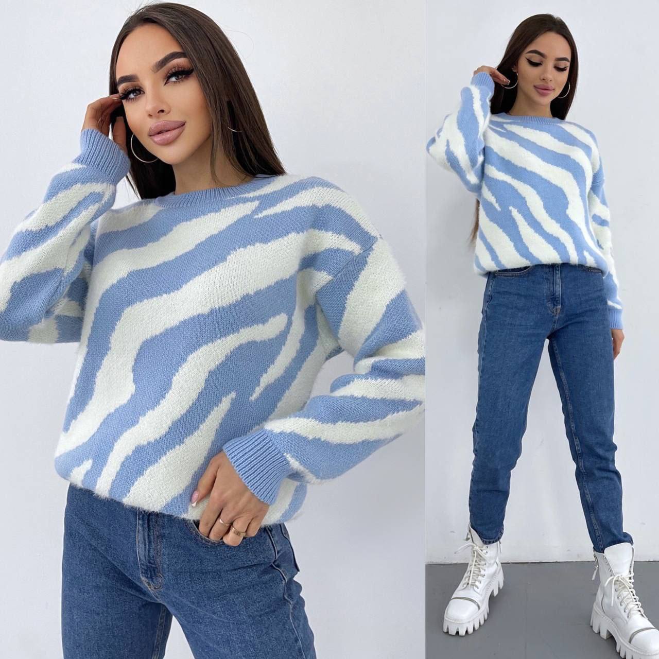 Woman wearing a blue and white zebra pattern sweater with jeans and white boots on a plain background