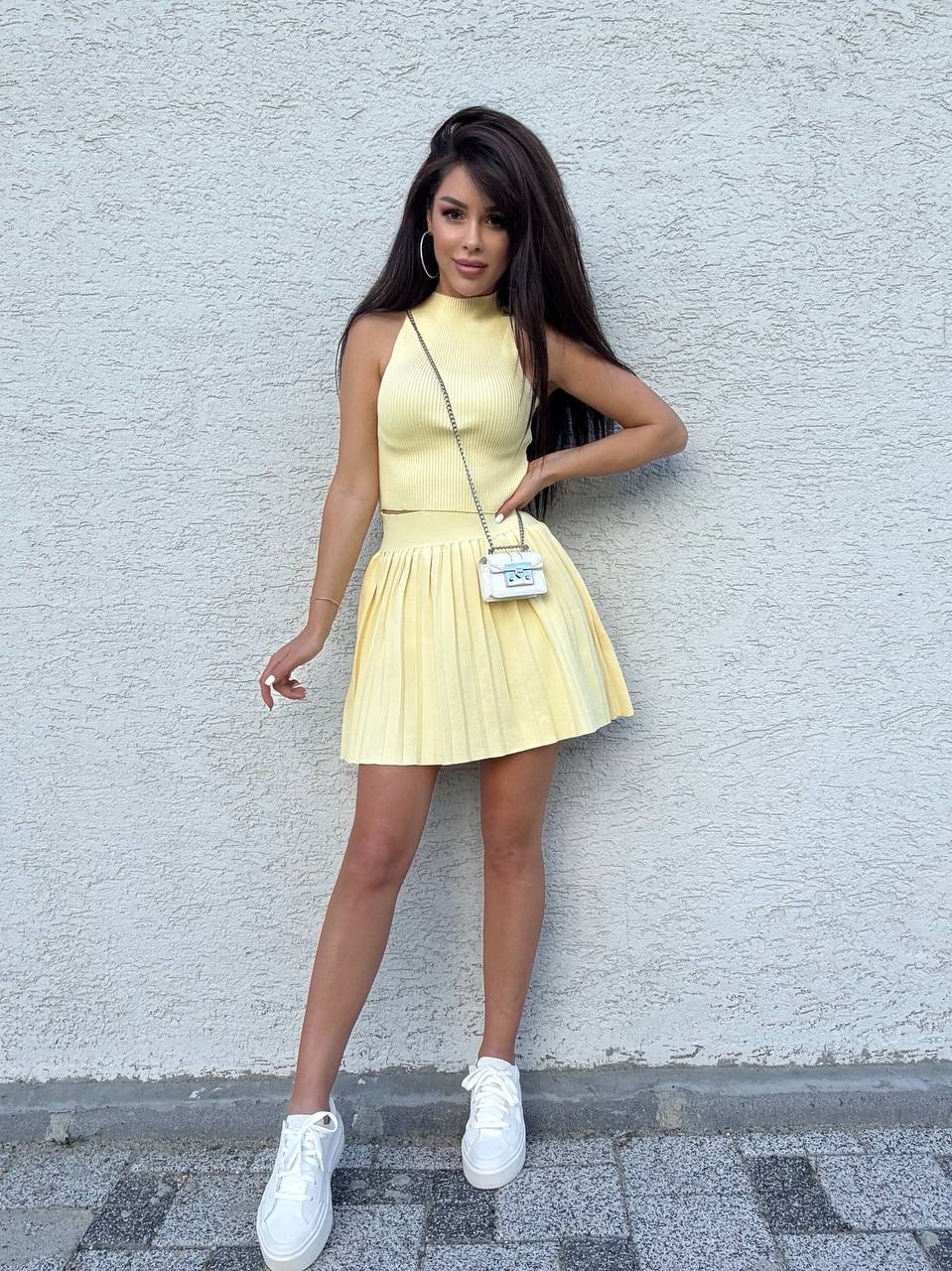 Woman wearing a yellow dress with pleated skirt against a light gray wall.