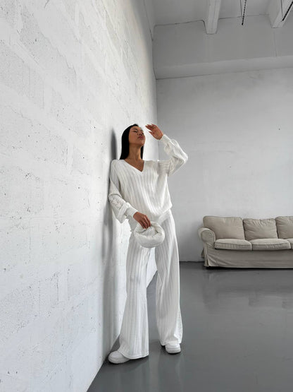 Woman in a white outfit standing against a white wall in a minimalistic room with a gray sofa.