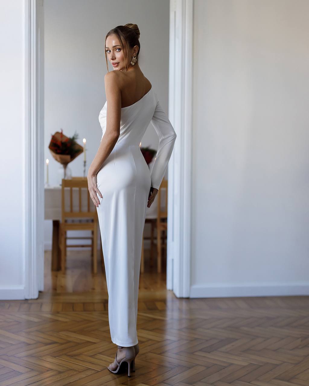 Woman in a white one-shoulder dress standing in a room with wooden flooring.