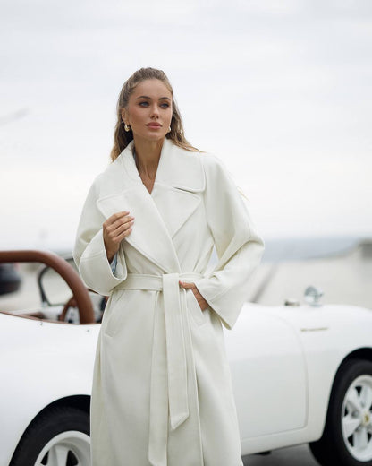 Woman in a white coat standing next to a vintage car with a blurred background