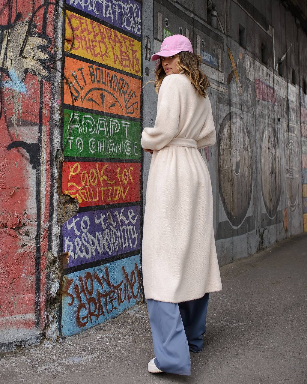 Woman in a long coat and pink cap standing next to a colorful mural with motivational text on a wall.