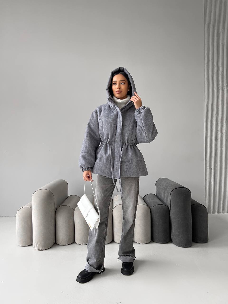 Person wearing a gray hooded jacket and pants standing against a plain background with gray abstract sculptures.