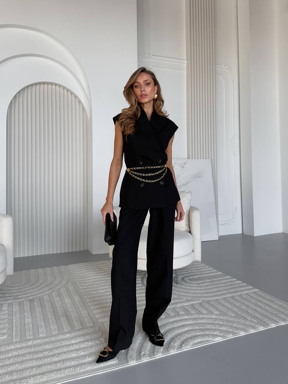 Woman in a black outfit with gold accents standing in a modern interior setting.