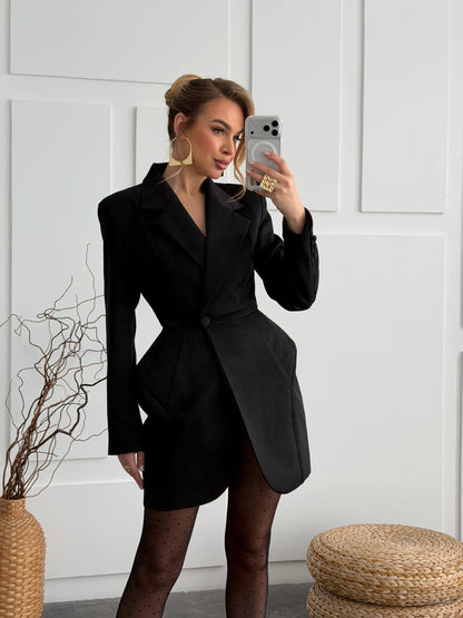 A woman takes a mirror selfie wearing a black tailored blazer-style mini dress with structured shoulders, a wrap-style front, and a cinched waist. She is wearing sheer black tights and large gold hoop earrings, standing against a white paneled wall with decorative dried branches in a woven vase and a round woven stool nearby.