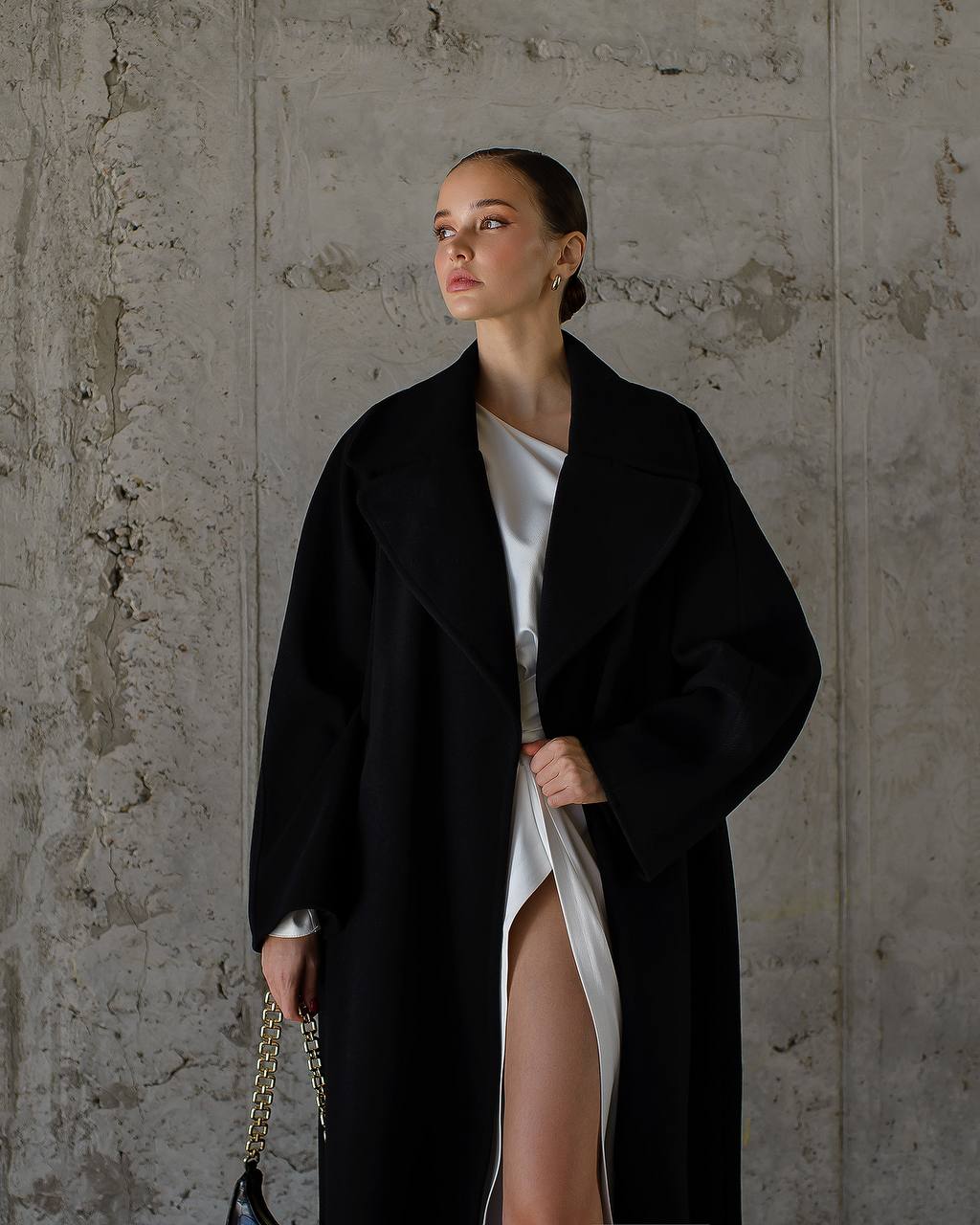 Woman wearing a black coat over a white dress against a textured wall.