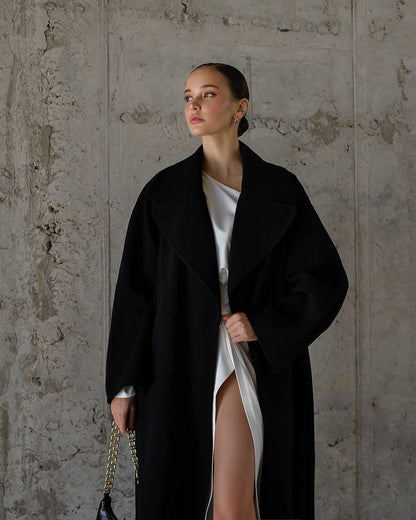 Woman wearing a black coat over a white dress against a textured wall.