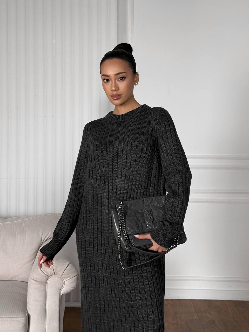 Woman in a black outfit holding a black handbag in a room with a white wall and gray sofa.