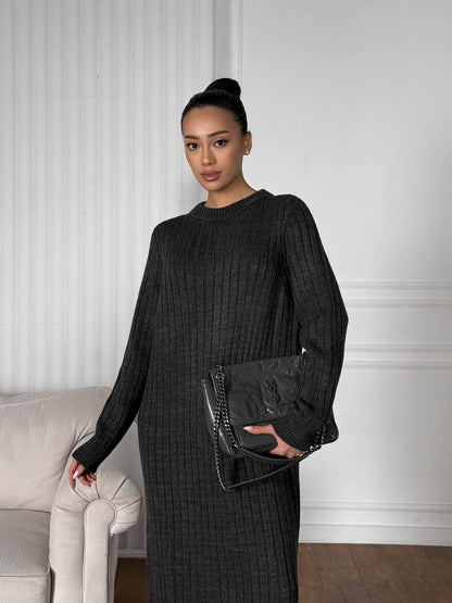 Woman in a black outfit holding a black handbag in a room with a white wall and gray sofa.