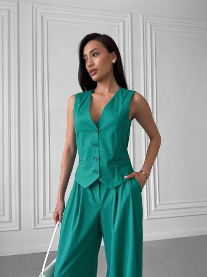 Woman wearing a green vest and pants set against a white paneled wall.