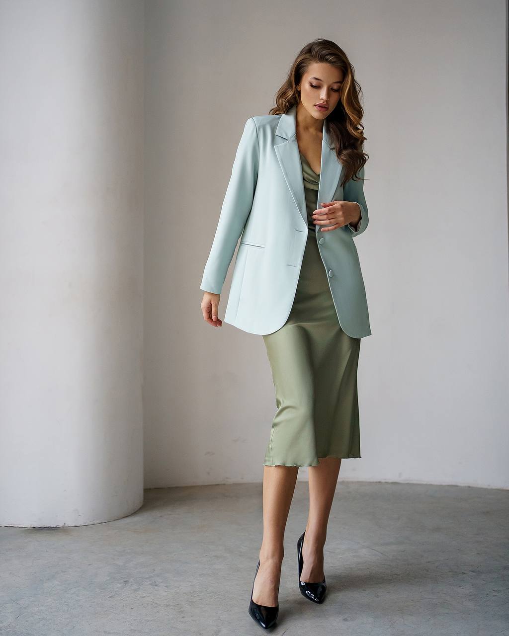 A woman models a light blue tailored blazer over an olive green satin midi dress, paired with black pointed-toe heels, standing in a minimalist studio with a white column and gray floor.
