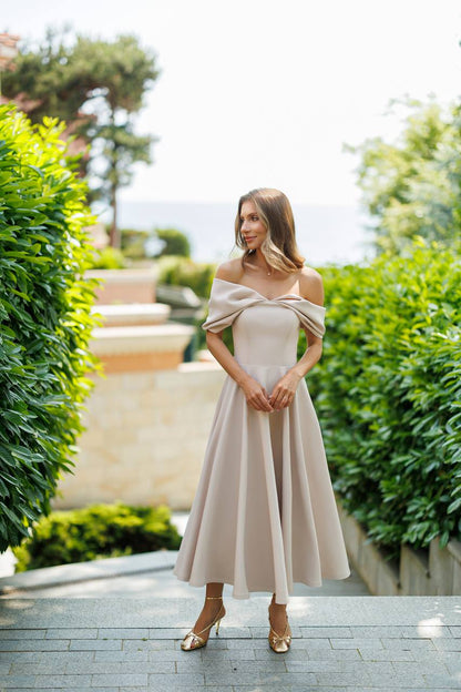 Woman in a beige off-shoulder dress standing outdoors with greenery and a path in the background