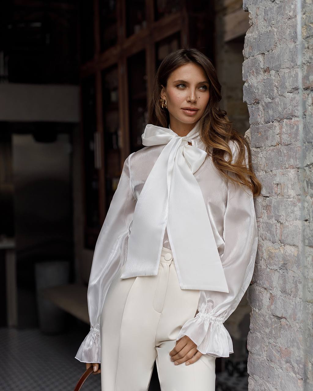 Woman wearing a white blouse with a large bow tie standing against a stone wall.