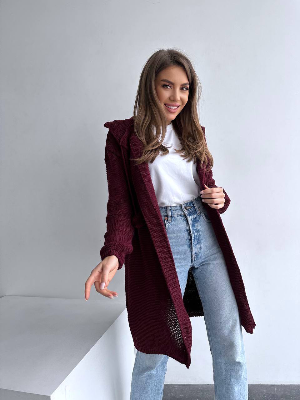 Woman wearing a burgundy coat, white shirt, and light blue jeans against a plain background