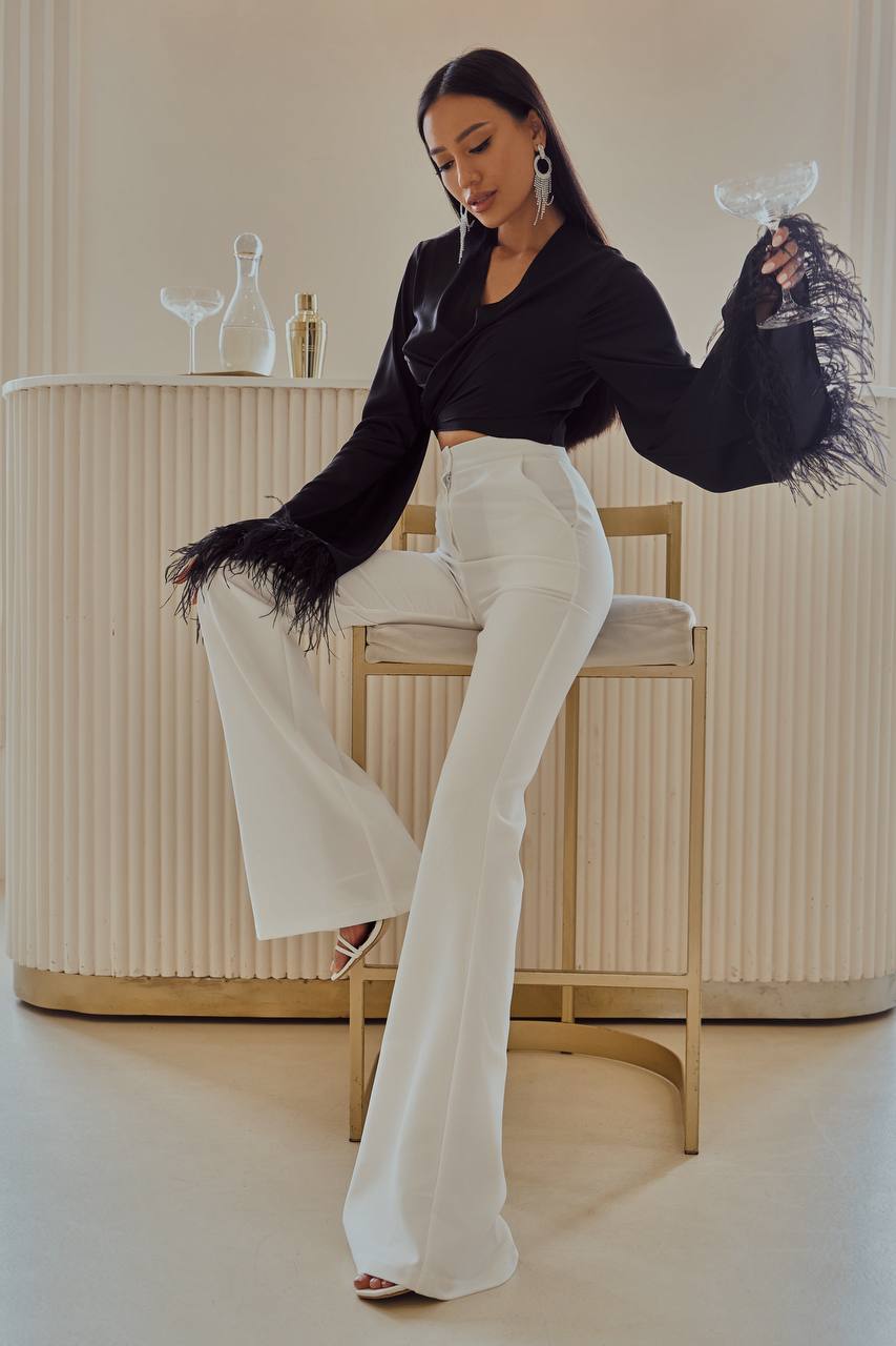 Woman in black top with fringed sleeves and white pants posing in a minimalistic room.