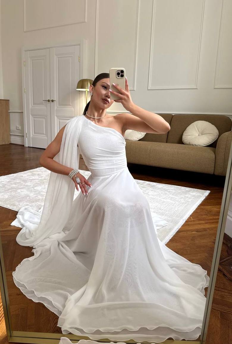 Woman in a white one-shoulder dress taking a mirror selfie in a room with a couch and rug.