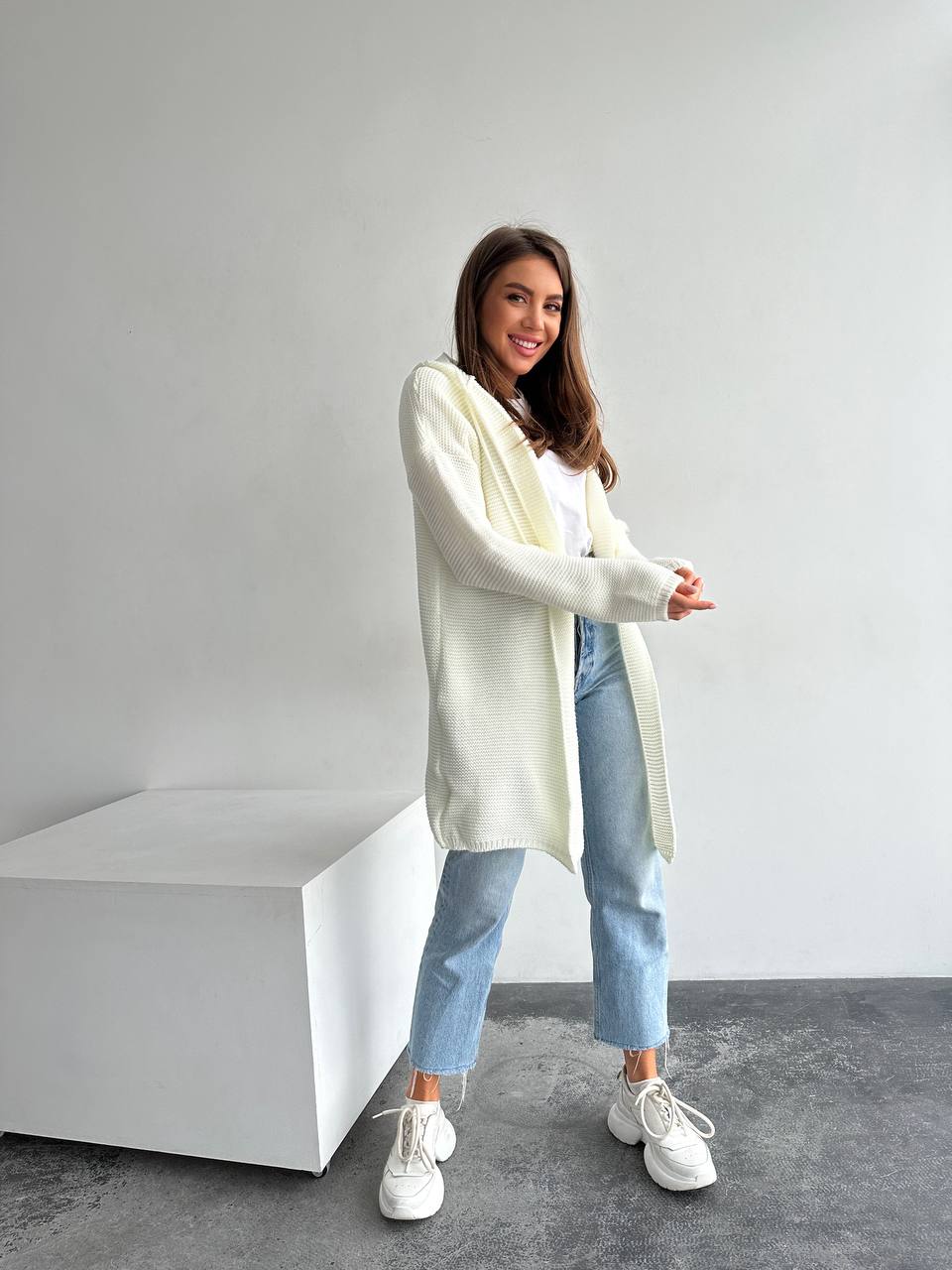 Woman wearing a long cream cardigan, light blue jeans, and white sneakers in a minimalistic setting.