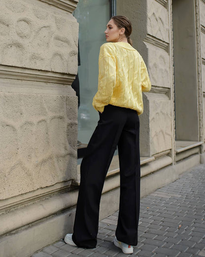 Woman wearing a yellow jacket and black pants standing against a stone building.