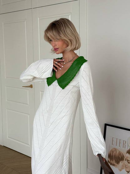 Woman wearing a white dress with a green neckline against a white wall.