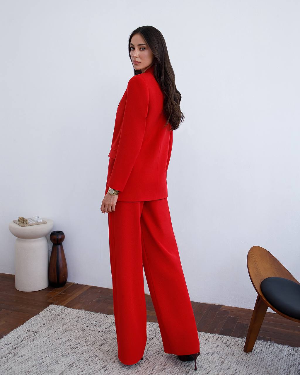 Woman wearing a red suit standing in a room with a white wall and wooden furniture.