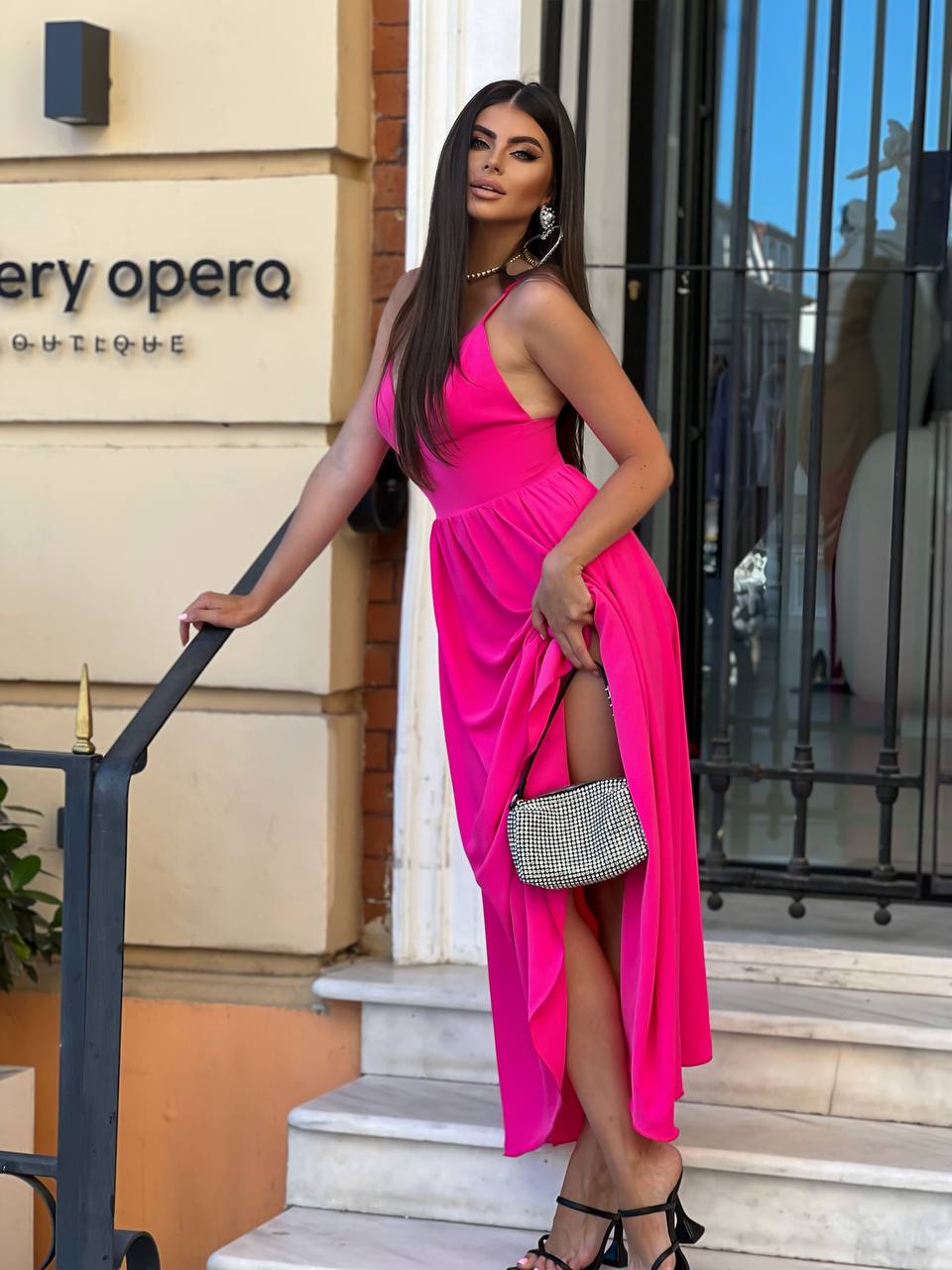A woman stands on outdoor stone steps in front of a boutique building, wearing a bright hot pink halter-neck midi dress with a thigh-high slit and holding a small silver handbag.