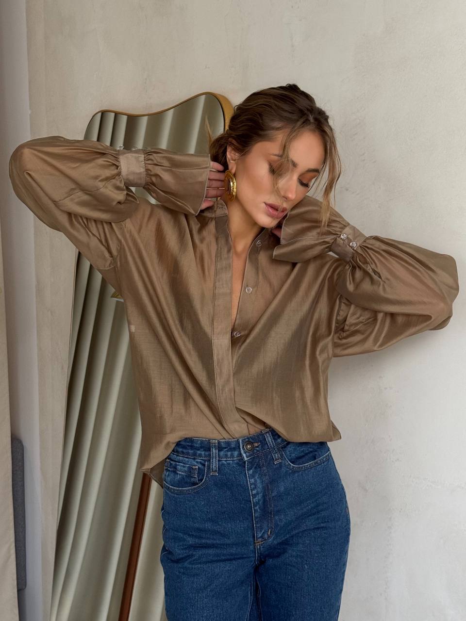 Woman wearing a brown satin shirt and blue jeans against a neutral wall.