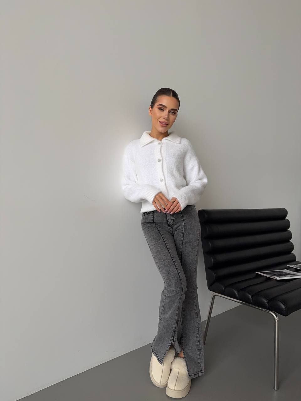 Woman in a white sweater and gray pants standing next to a black chair against a plain wall.