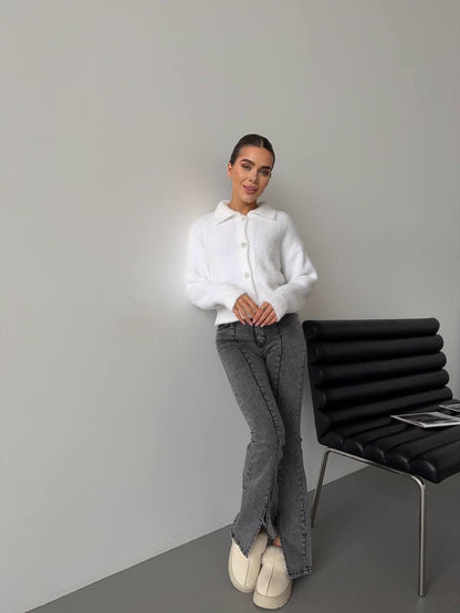 Woman in a white sweater and gray pants standing next to a black chair against a plain wall.