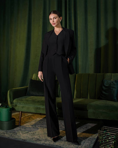 Woman in a black suit standing in a room with green curtains and a green sofa.