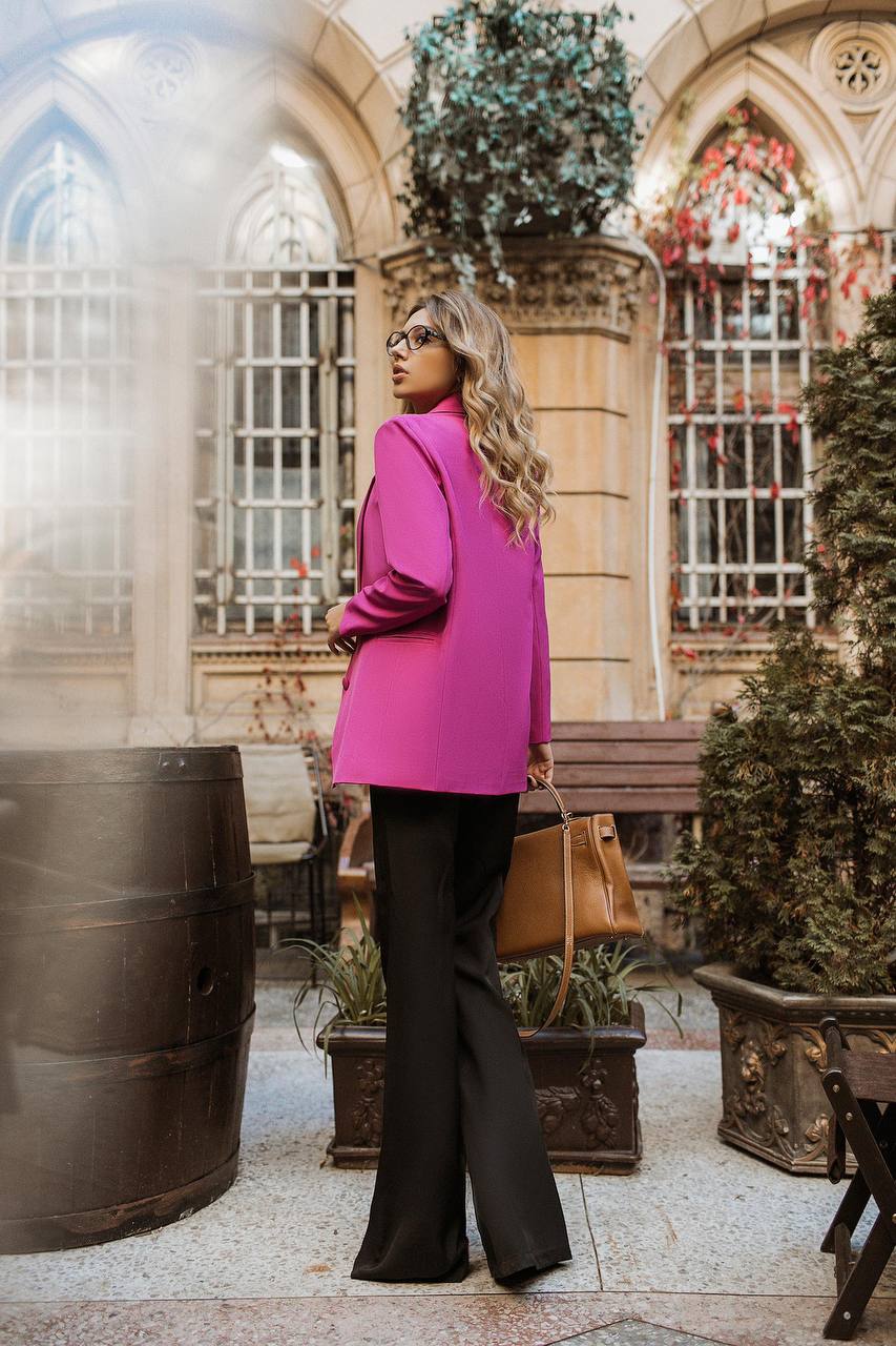 Woman in a pink blazer and black pants walking outdoors with decorative plants and architecture in the background.