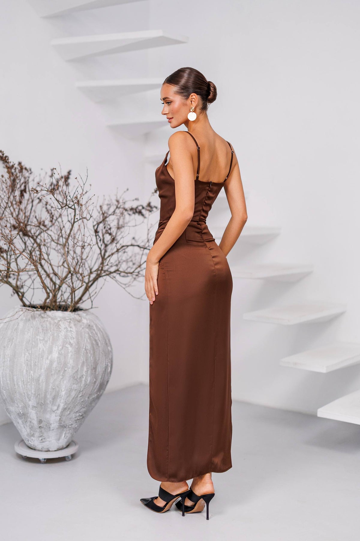 Woman in a brown dress standing in a modern indoor setting with a white vase and plant.
