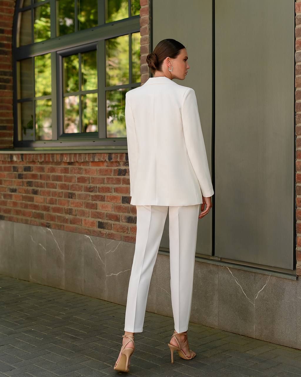 Woman in a white suit standing against a brick wall with a window.