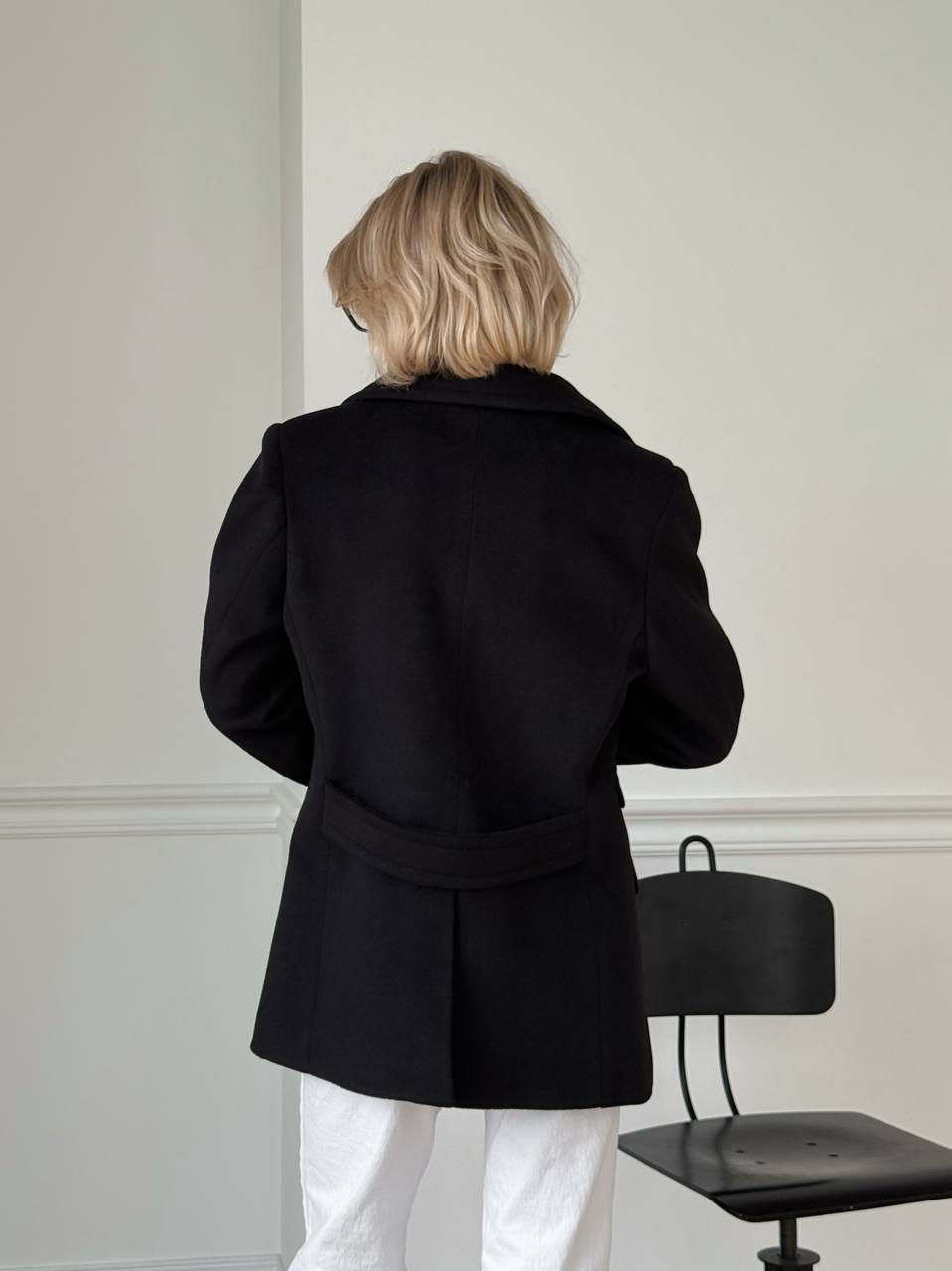 Person wearing a black coat in a minimalistic room with a chair.