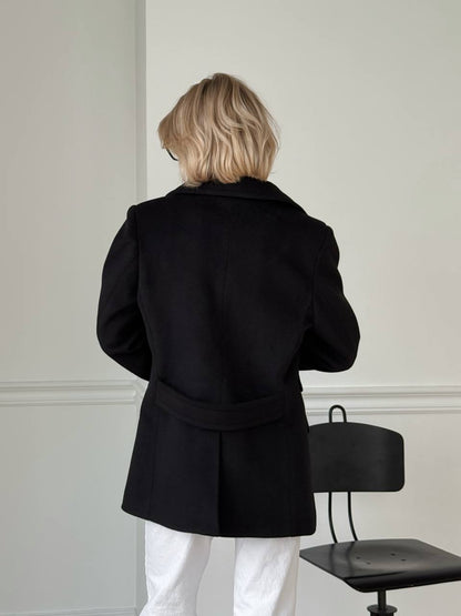 Person wearing a black coat in a minimalistic room with a chair.