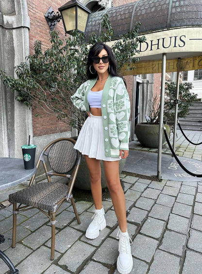 Woman in a light green cardigan and white skirt standing outdoors near a building entrance.