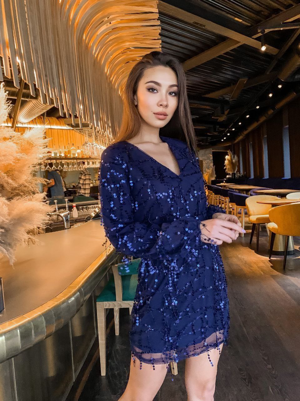 Woman in a blue sequin dress standing in a modern indoor setting with decorative elements.
