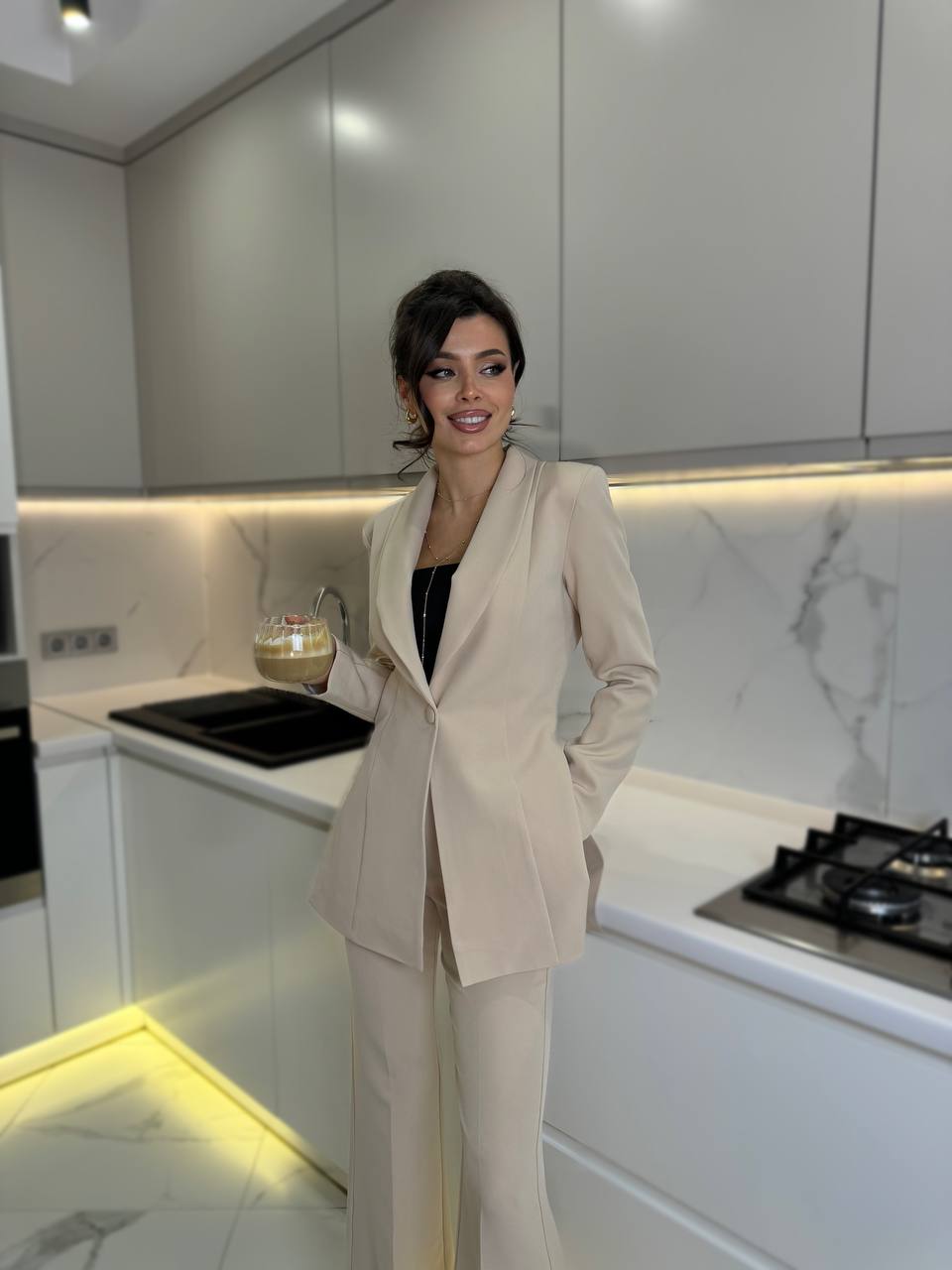 Woman in a beige suit standing in a modern kitchen with white cabinets and marble countertop.