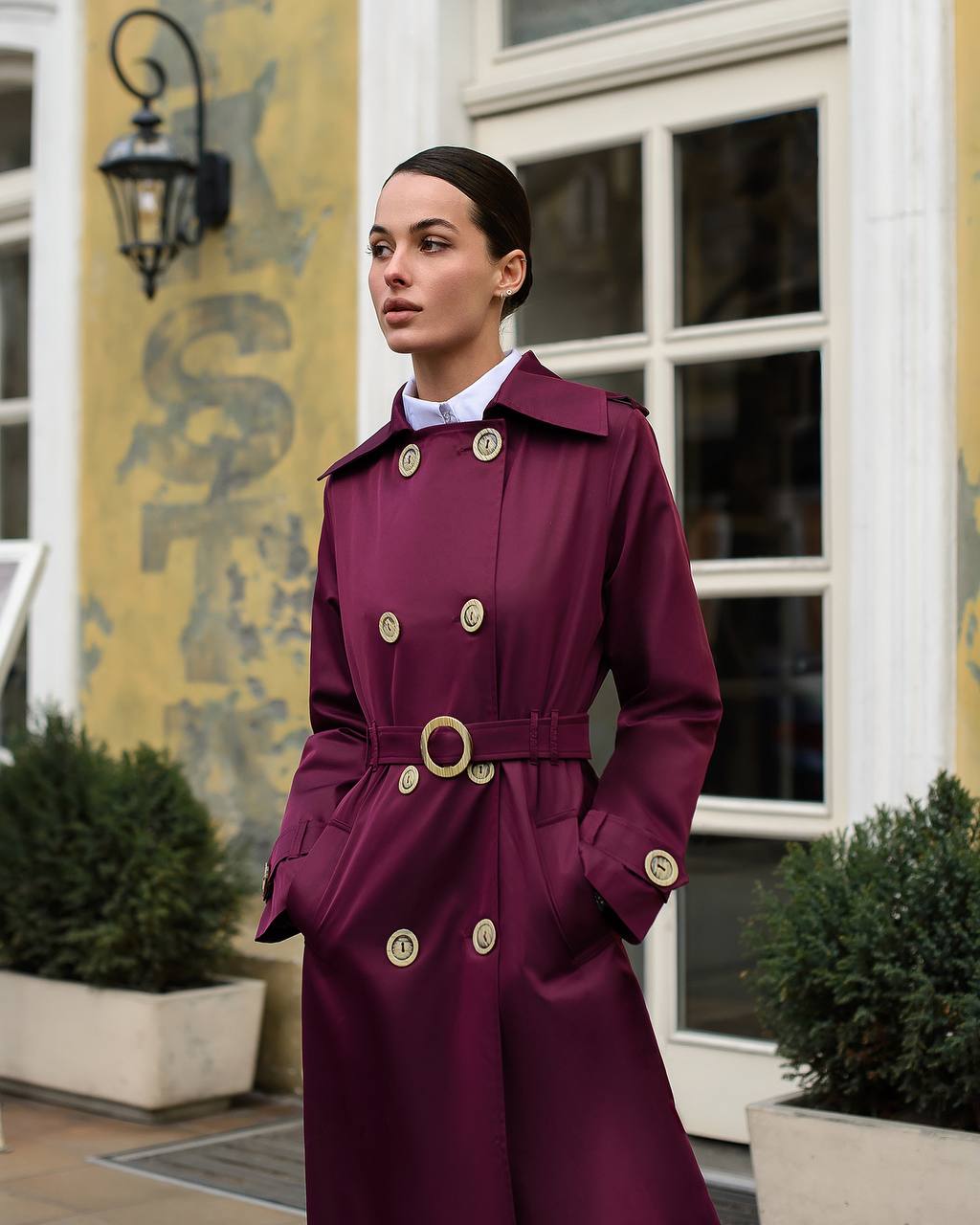 Woman wearing a burgundy trench coat with gold buttons in an urban setting.