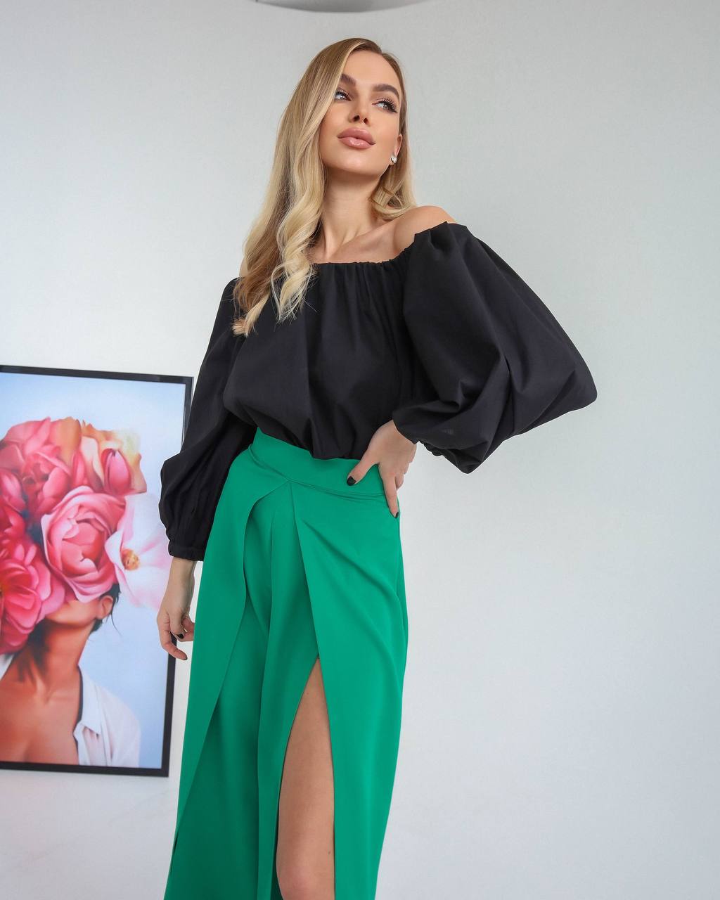 Woman wearing a black off-shoulder top and green skirt in a room with a colorful painting on the wall.