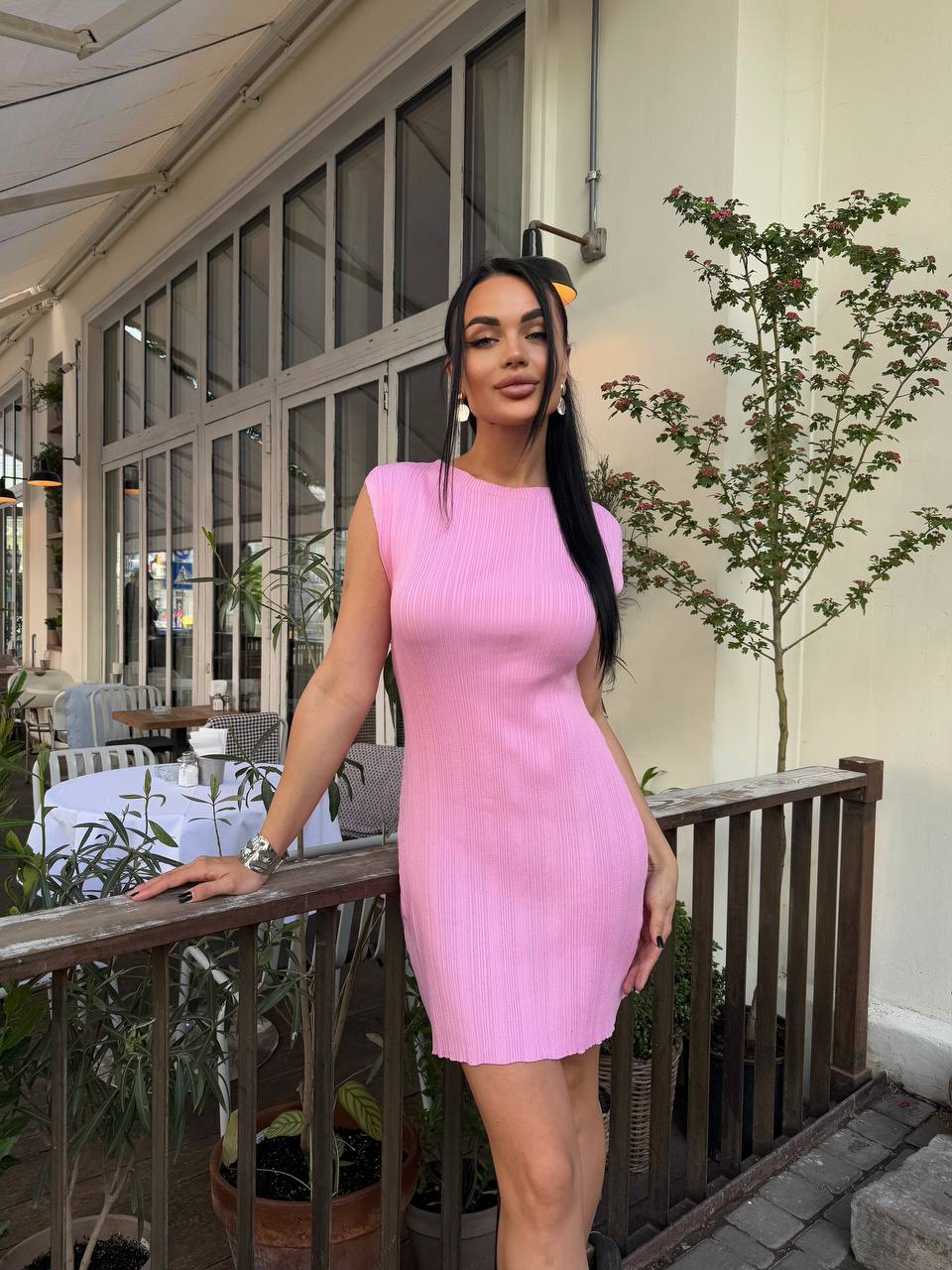 Woman in a pink dress standing on a balcony with plants and tables in the background
