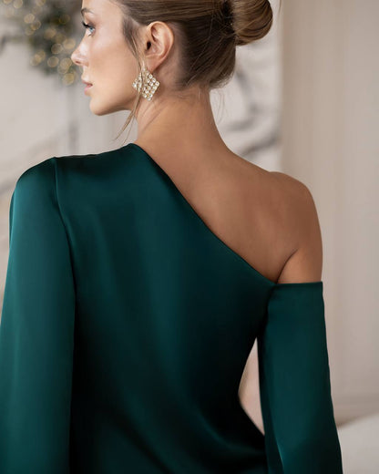 A woman shown from the back wearing a dark emerald green one-shoulder dress with a long draped sleeve and a high neckline. She is also wearing a large, textured gold statement earring. The background is a softly blurred indoor setting with warm neutral tones.
