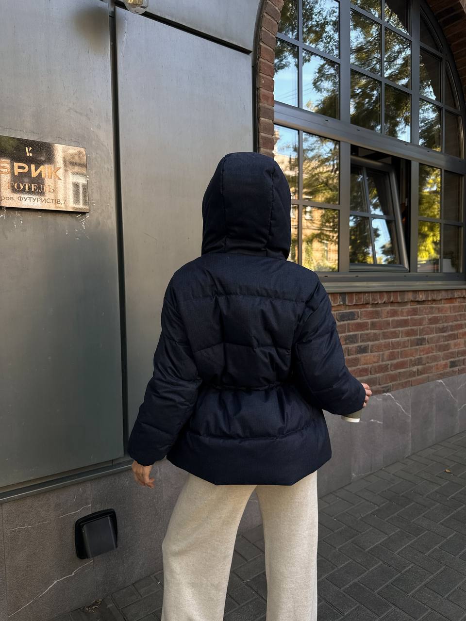 Person wearing a black puffer jacket standing outside a building with a sign.