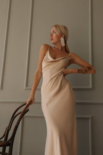 Woman in a beige dress standing against a neutral wall.