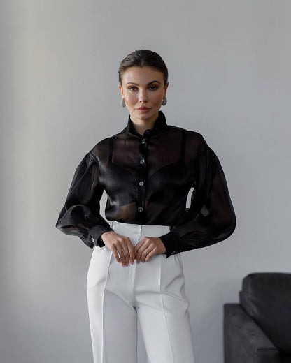 Woman wearing a black sheer blouse and white pants against a gray background