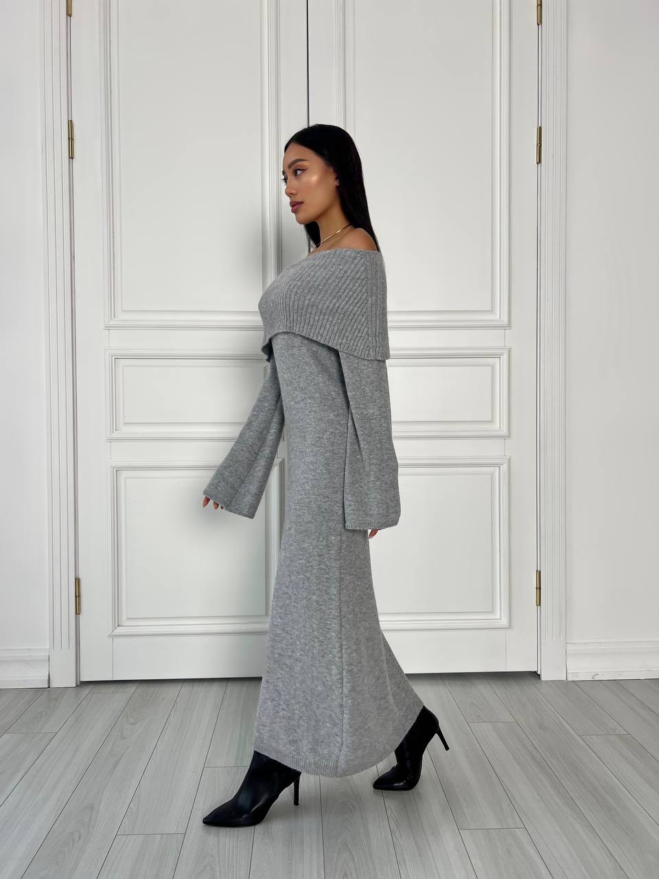 Woman wearing a gray off-shoulder dress against a white paneled wall.