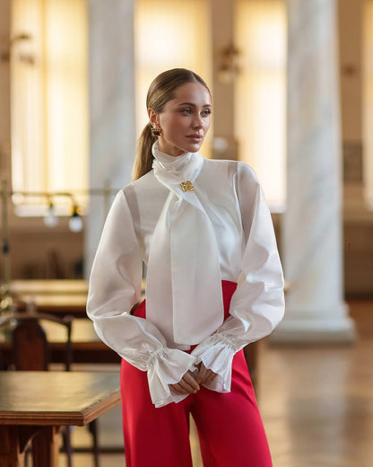 Woman wearing a white blouse with a high collar and red pants in an indoor setting.