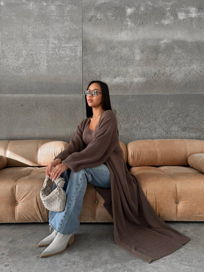 Woman sitting on a brown sofa wearing sunglasses and holding a small bag, with a gray wall in the background.